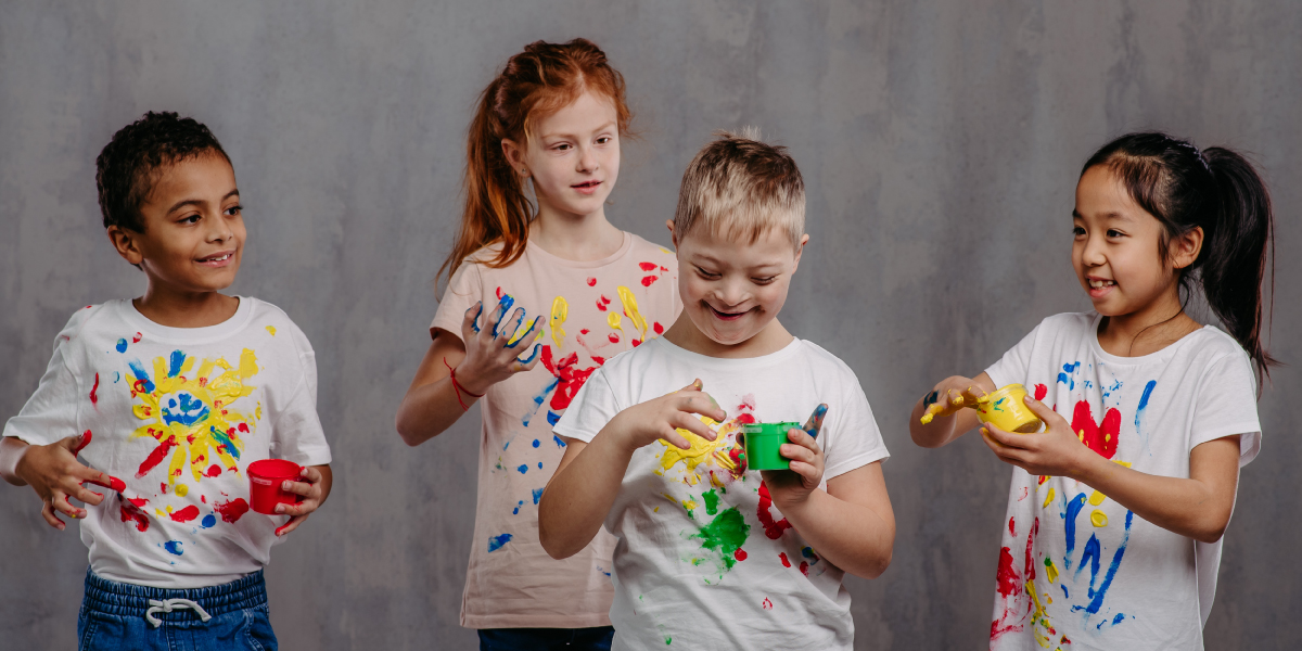 A diverse group of four smiling children wearing white tee shirts are finger painting. They are each holding a cup of paint and have painted designs on their shirts.