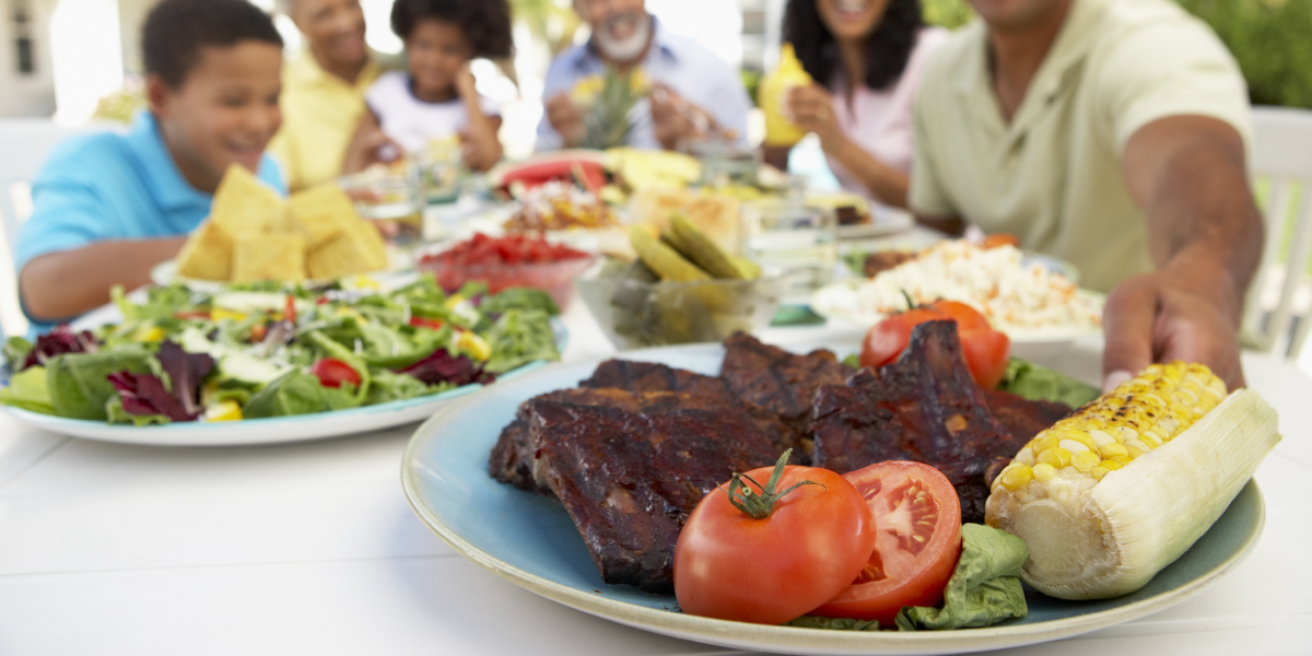 Multigenerational family enjoying eating outside with a table full of plates of food. The focus in on a plate in the foreground. The ceramic plate holds barbecued meat, roasted corn on the cob, fresh sliced tomatoes and lettuce. A plate of salad and a bowl of pickles can be seen in semi-focus.