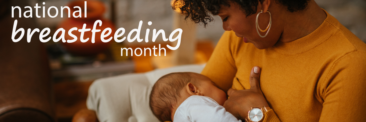 Woman with dark skin and curly black hair is breastfeeding her infant. She sits in a comfortable living space, with low lamp light, wearing an orange top, gold earrings, and a gold watch. She smiling at the infant at her breast.