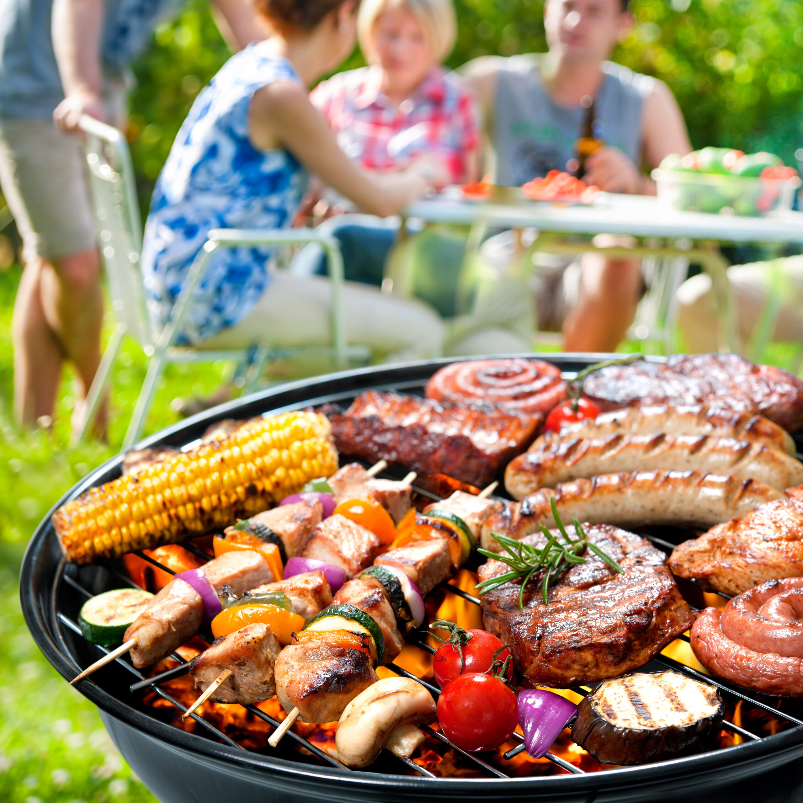 Meats and vegetables grilling over red-hot coals on outdoor uncovered grill. Foreground focus is on a round outdoor grill loaded with a variety of meats and vegetables cooking over red-hot coals. A group of friends congregate at an outdoor table in bright sunlight in the soft-focus background.