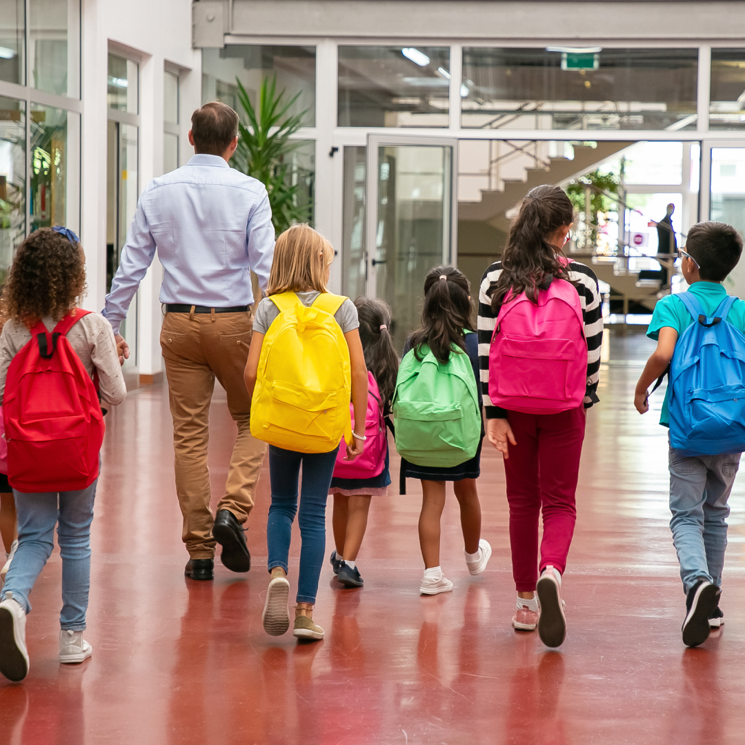 Back to School group Seen from the back, a diverse group of elementary age children in colorful clothes and backpacks are being led by a male-presenting adult through a brightly lit school corridor.